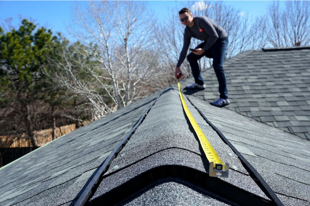 A handyman on top of a roof uses yellow measuring tape to measure the size of the roof