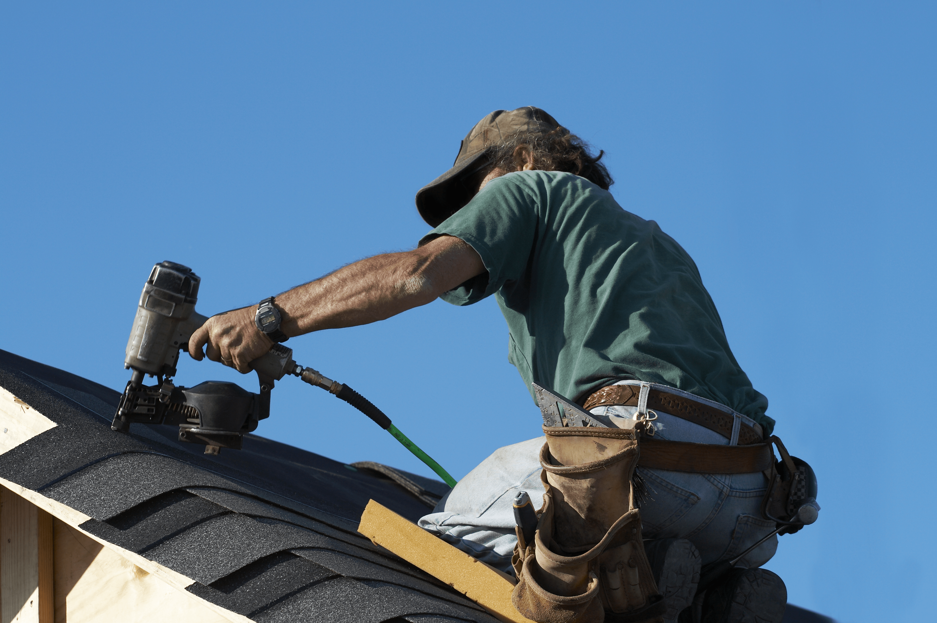 The back of a handyman kneeling on a roof doing roof repair on a sunny day.