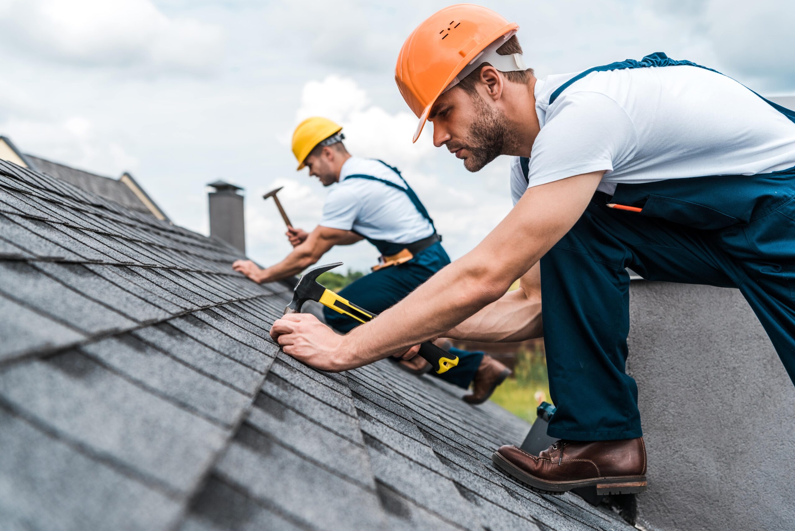 shutterstock_1479491051-min A handyman and his partner stand on a roof and use hammers to do their jobs.